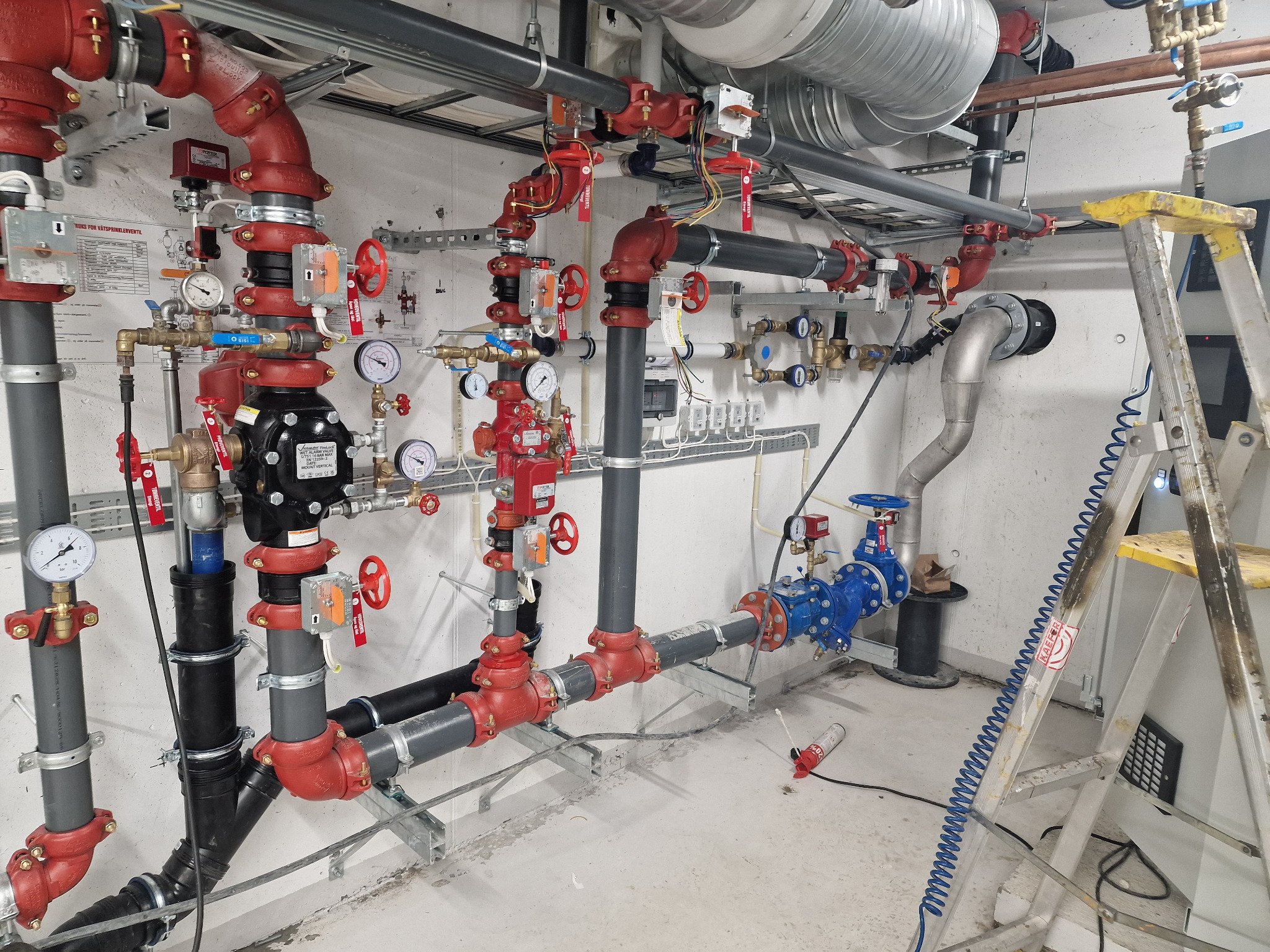 Złożony system rur w kolorach czerwonym, szarym i czarnym, z zaworami, manometrami i przewodami elektrycznymi, zamontowany na białej ścianie, widoczna drabina aluminiowa z żółtym stopniem.