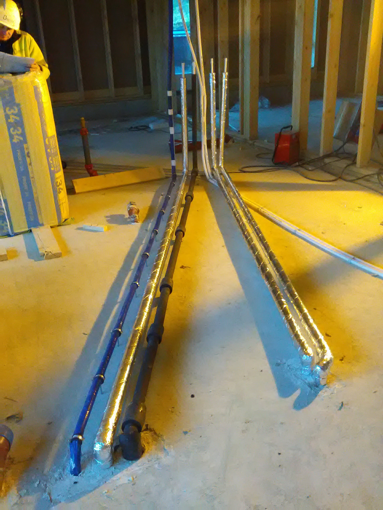 Instalacja rur wodnych i grzewczych w surowym stanie, rury częściowo zaizolowane folią, widoczne drewniane elementy konstrukcji budynku oraz materiały izolacyjne