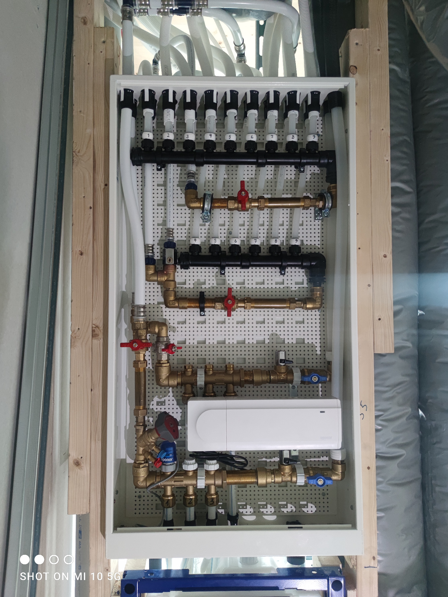 Rozdzielacz ogrzewania podłogowego w skrzynce montażowej, widoczne rury doprowadzające i odprowadzające wodę, zawory regulacyjne i elektroniczny sterownik, całość zamontowana na drewnianej...