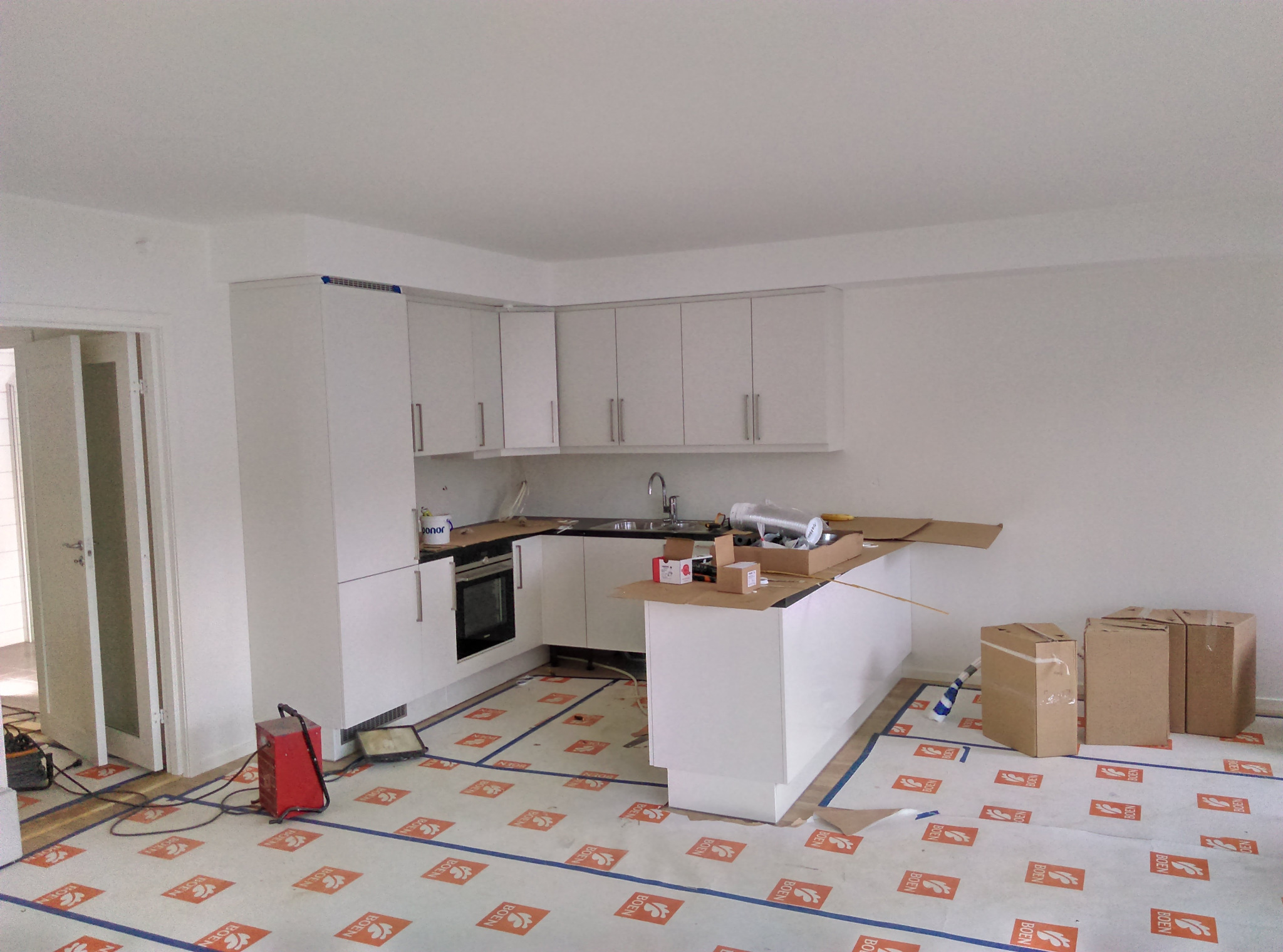 Montaż białej zabudowy kuchennej w trakcie prac wykończeniowych, widoczne kartony, narzędzia i folia ochronna na podłodze z logo BOEN.