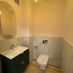 Remonty SAV-REM - Wykończona łazienka z toaletą, umywalką nablatową na szafce z frezowanymi frontami w kolorze ciemnej zieleni, lustrem w owalnej ramie i marmurowymi płytkami do połowy ściany.