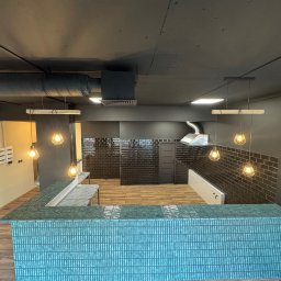 Remonty SAV-REM - Wykończone wnętrze biurowe w Warszawie z turkusową ladą recepcyjną z płytek, czarną ścianą z płytek i nowoczesnym oświetleniem.