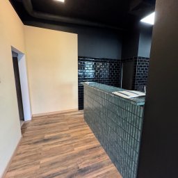 Remonty SAV-REM - Nowoczesne wnętrze z drewnopodobną podłogą, czarnym sufitem z widocznymi rurami, kremową ścianą i ladą wykończoną drobną, turkusową mozaiką, za którą widać czarne płytki ułożone w cegiełkę.