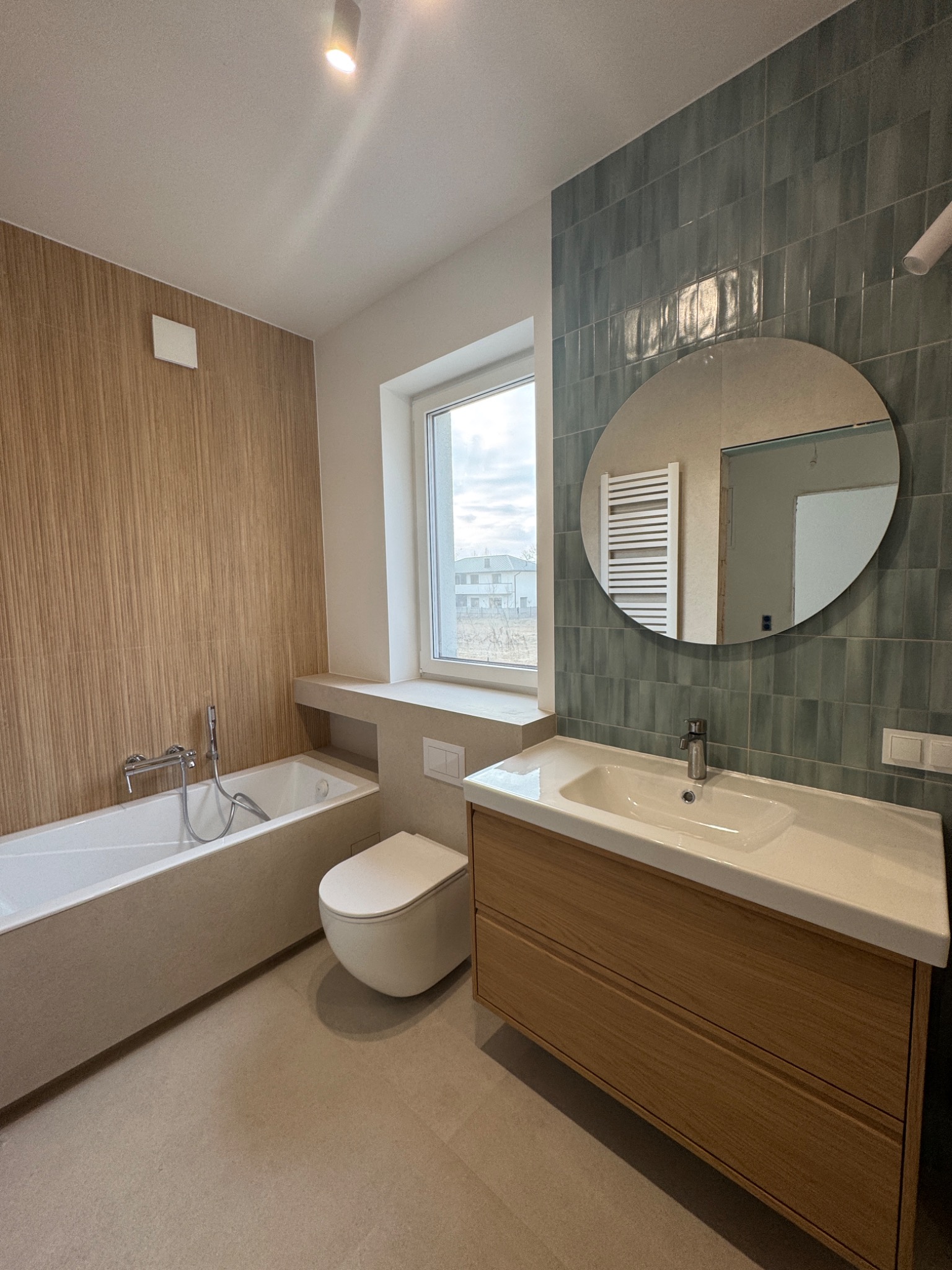 Nowoczesna łazienka z drewnianą zabudową umywalki, okrągłym lustrem i pionowymi turkusowymi płytkami na ścianie, widok na wannę i podwieszaną toaletę.