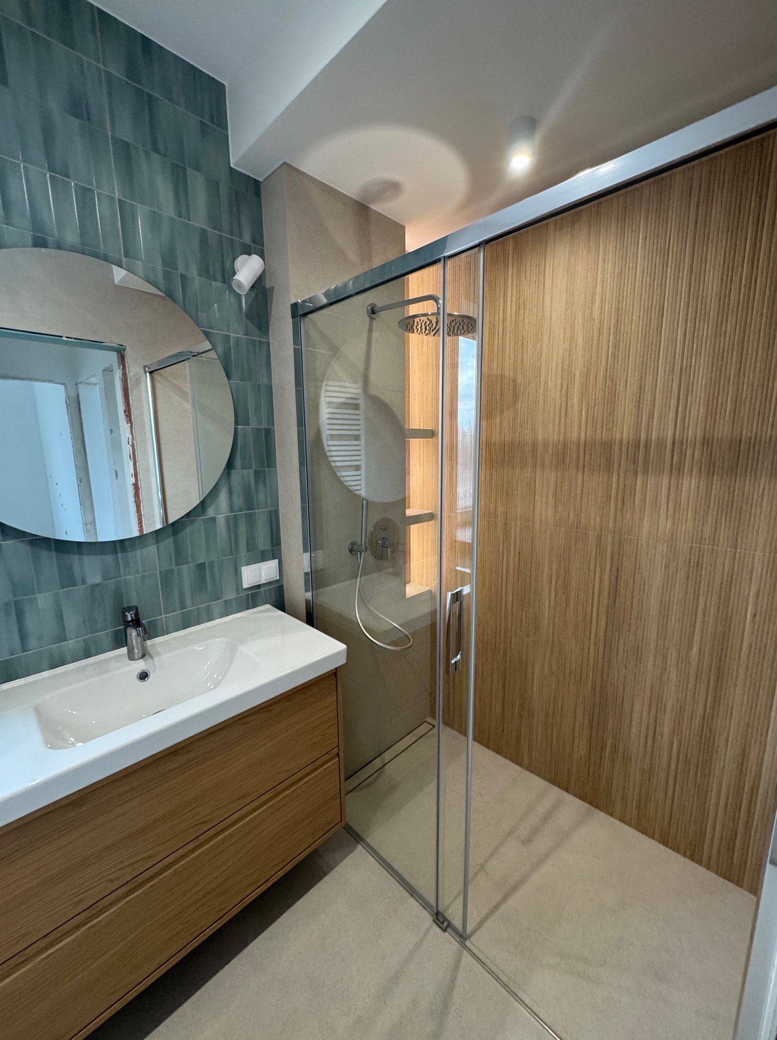 Nowoczesna łazienka z kabiną prysznicową typu walk-in, umywalką nablatową na drewnianej szafce, okrągłym lustrem i turkusowymi płytkami na ścianie.