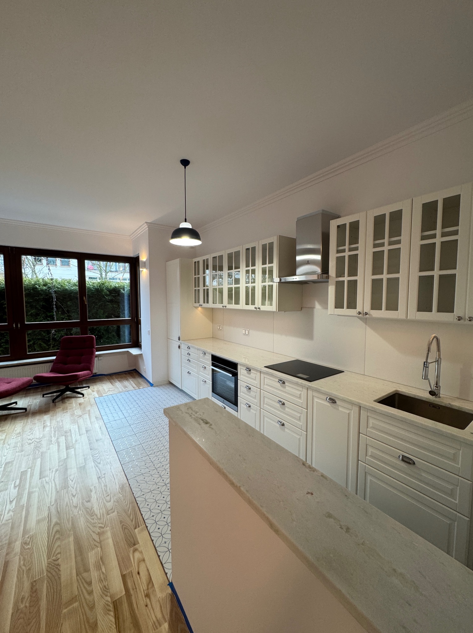 Wykończona kuchnia w stylu klasycznym z białymi szafkami, blatem z jasnego kamienia i drewnianą podłogą przechodzącą w geometryczne płytki. W tle widoczne okno z widokiem na zieleń i czerwony fotel.