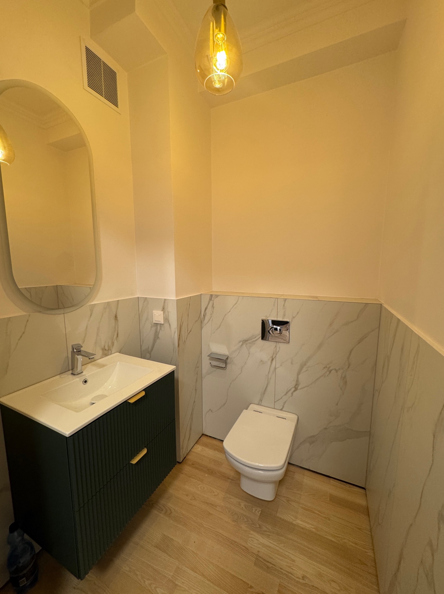 Wykończona łazienka z toaletą, umywalką nablatową na szafce z frezowanymi frontami w kolorze ciemnej zieleni, lustrem w owalnej ramie i marmurowymi płytkami do połowy ściany.