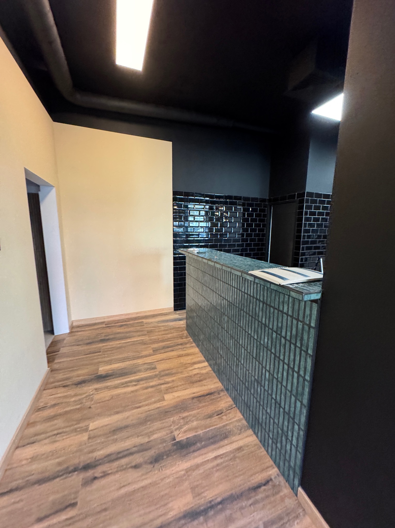 Nowoczesne wnętrze z drewnopodobną podłogą, czarnym sufitem z widocznymi rurami, kremową ścianą i ladą wykończoną drobną, turkusową mozaiką, za którą widać czarne płytki ułożone w cegiełkę.