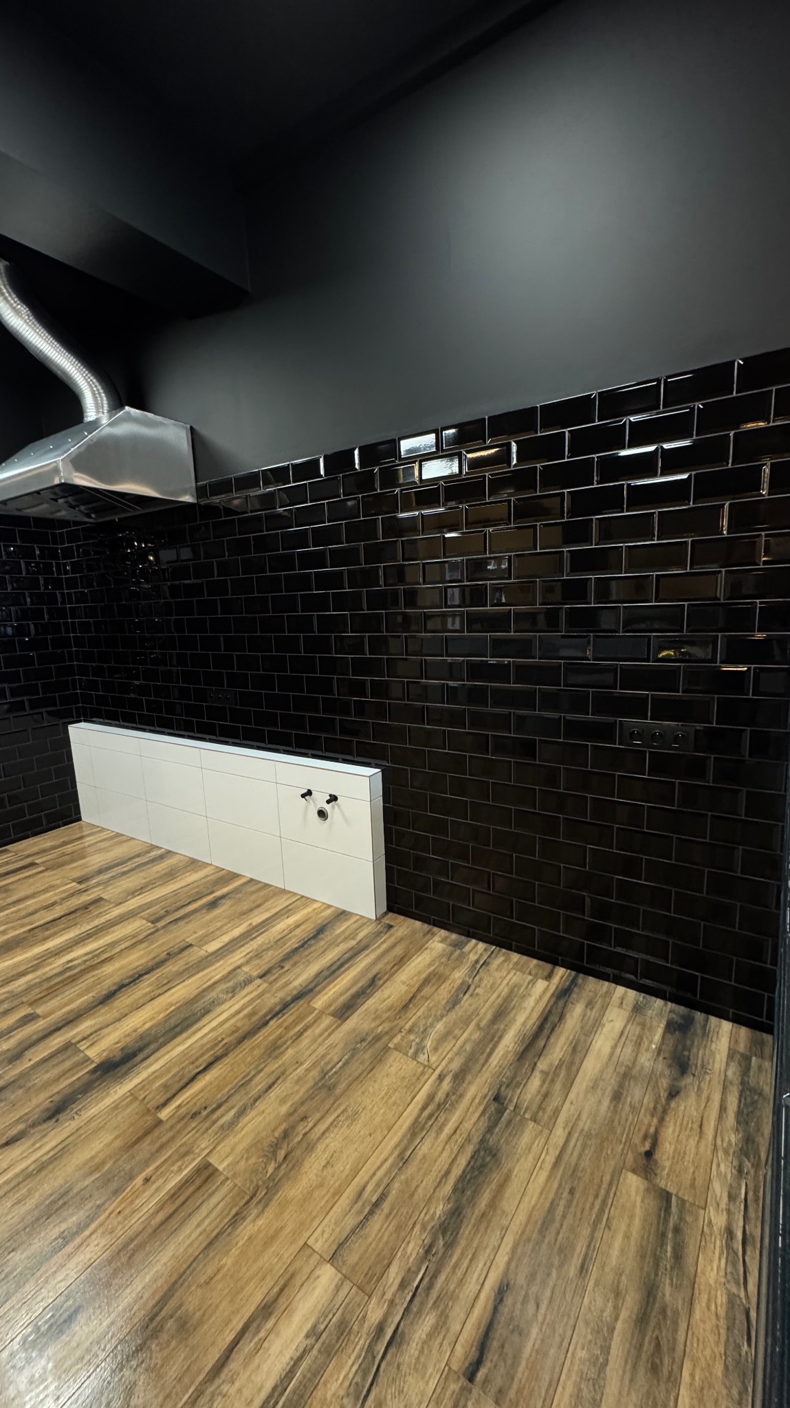 Nowoczesna łazienka z czarnymi, błyszczącymi płytkami typu metro na ścianach, drewnopodobną podłogą i białą zabudową pod umywalkę z widocznymi przyłączami wodnymi oraz metalowym okapem.