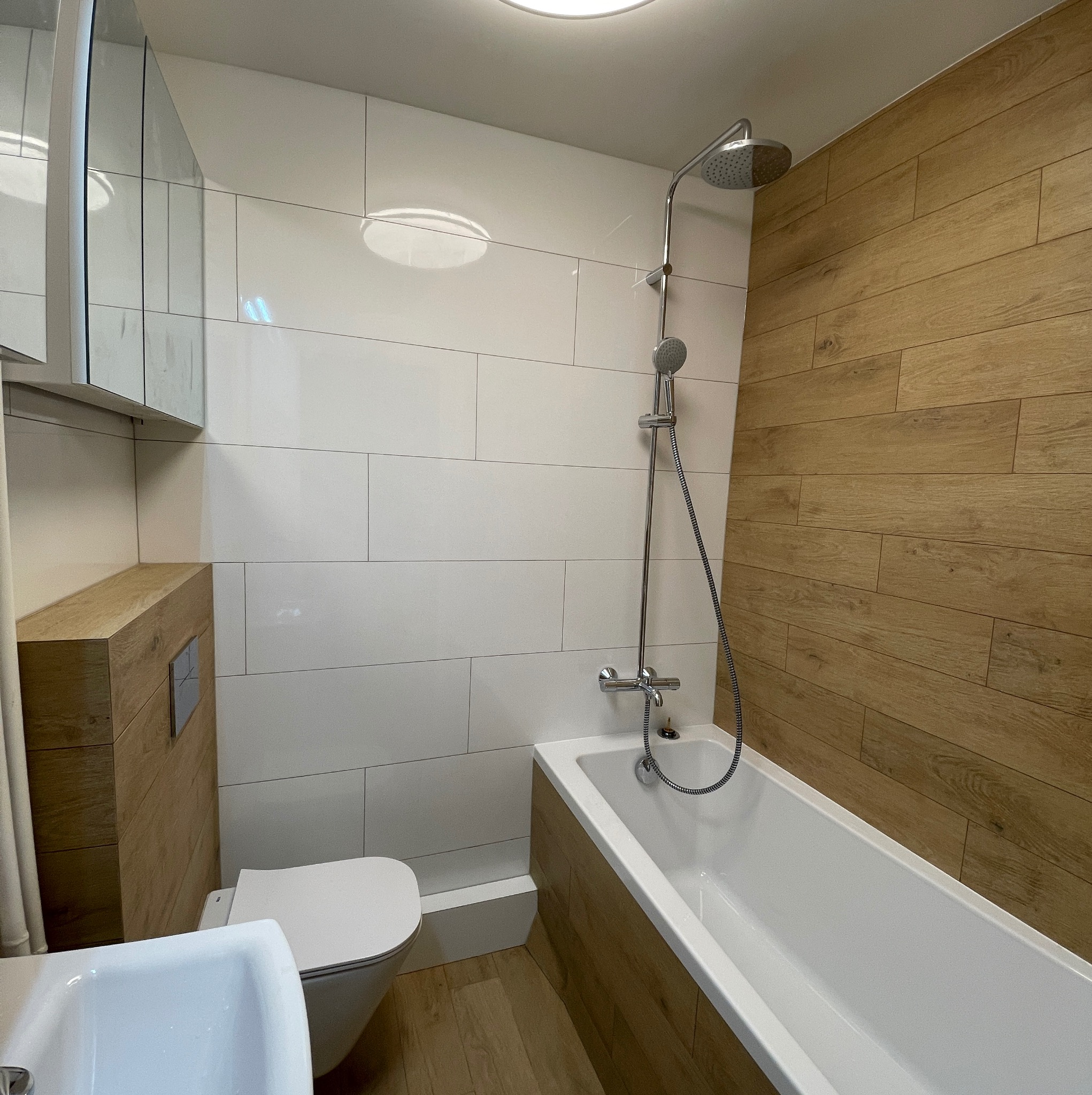 Nowoczesna łazienka z białymi płytkami, drewnopodobną okładziną i chromowaną armaturą, prezentująca minimalistyczny styl.