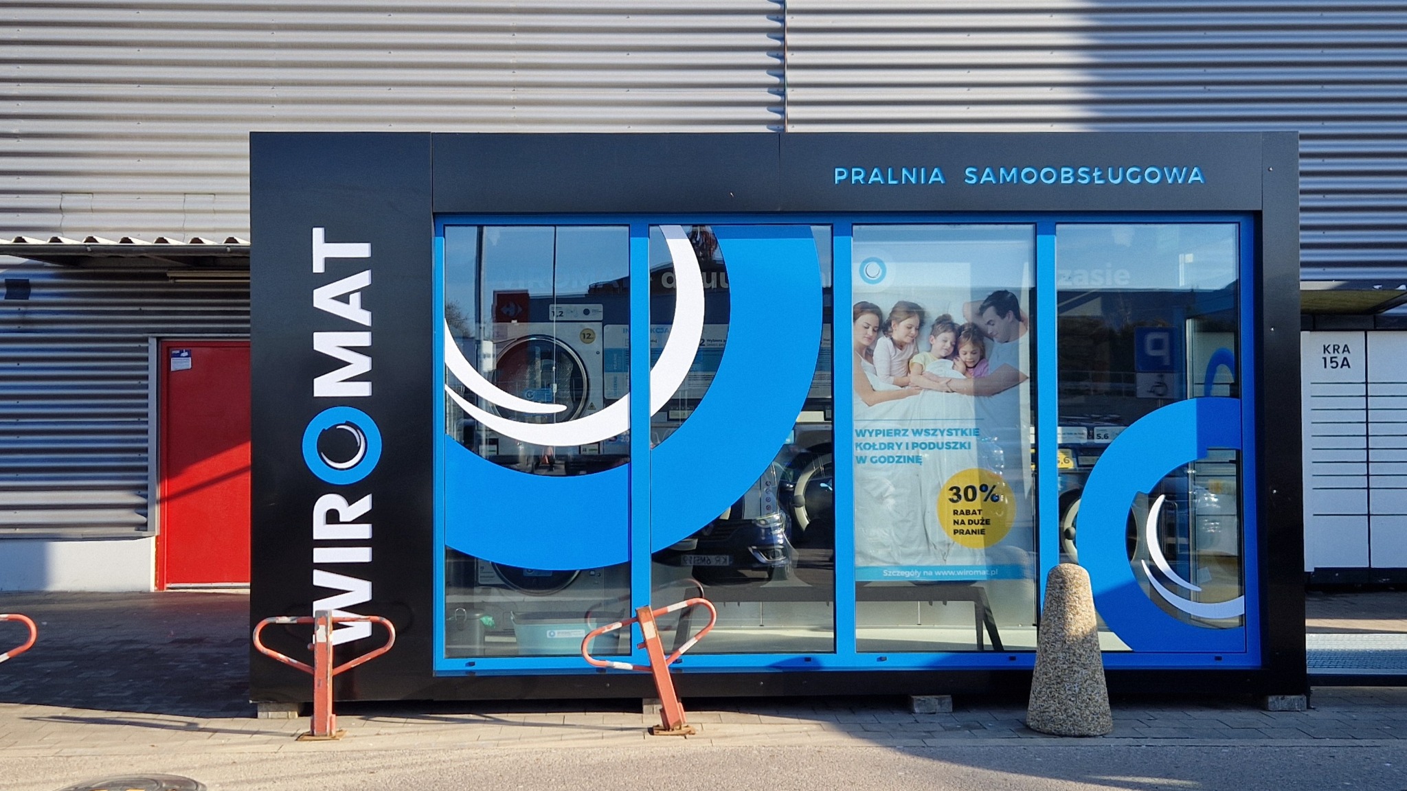 Zewnętrzny widok na samoobsługową pralnię w Krakowie, z widocznym logo 'Wiromat' i informacją o 30% rabacie na duże pranie. Za szybą widać pralki i plakat z uśmiechniętą rodziną.