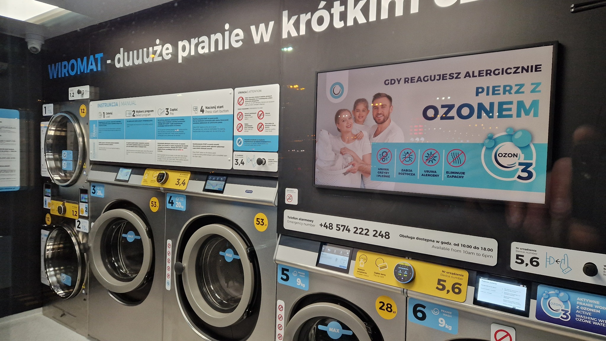 Wnętrze samoobsługowej pralni chemicznej z rzędem srebrnych pralek automatycznych, instrukcjami obsługi i reklamą prania z ozonem dla alergików.