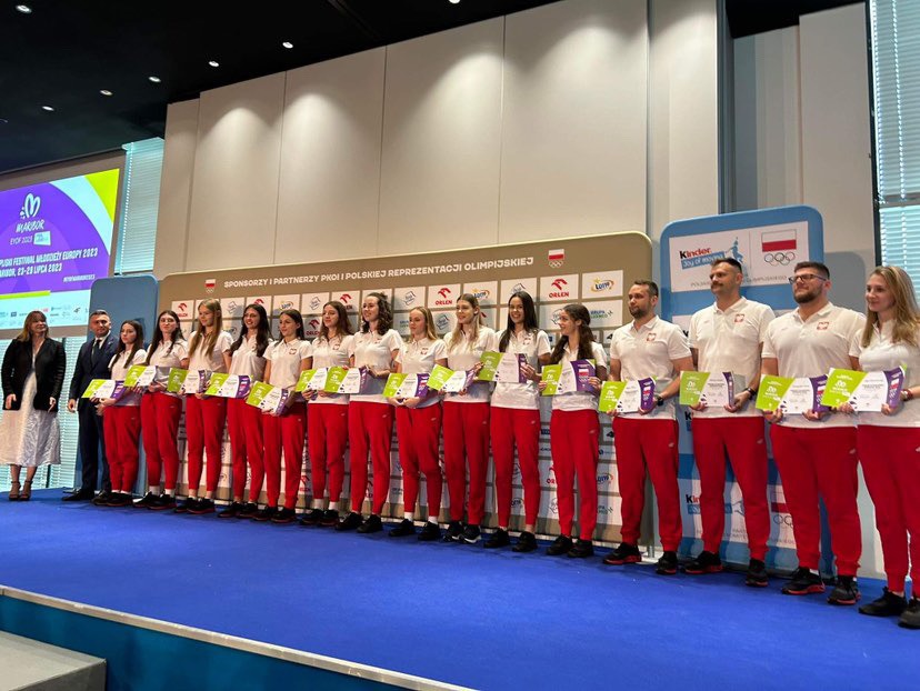Grupa sportowców reprezentacji Polski, w strojach sportowych z flagą, na scenie podczas ceremonii wręczenia nagród, trzymających certyfikaty na tle ścianki sponsorskiej.