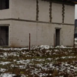 Surowy stan dwupiętrowego domu z pustaków, z widocznymi otworami okiennymi i drzwiowymi, oraz fragmentem dachu z czarnej dachówki, sfotografowany w pochmurny dzień na tle ziemi pokrytej resztkami...