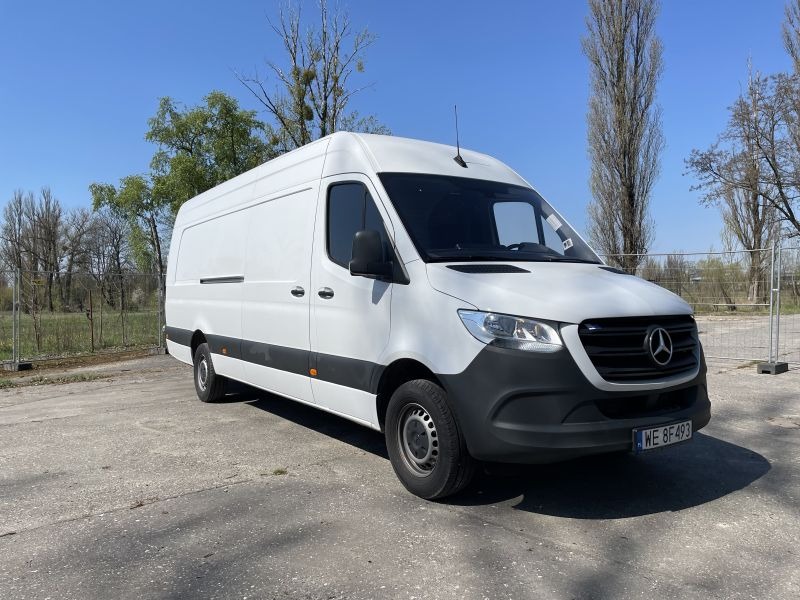 Biały samochód dostawczy Mercedes-Benz Sprinter na betonowym placu, widoczne drzewa w tle i błękitne niebo.