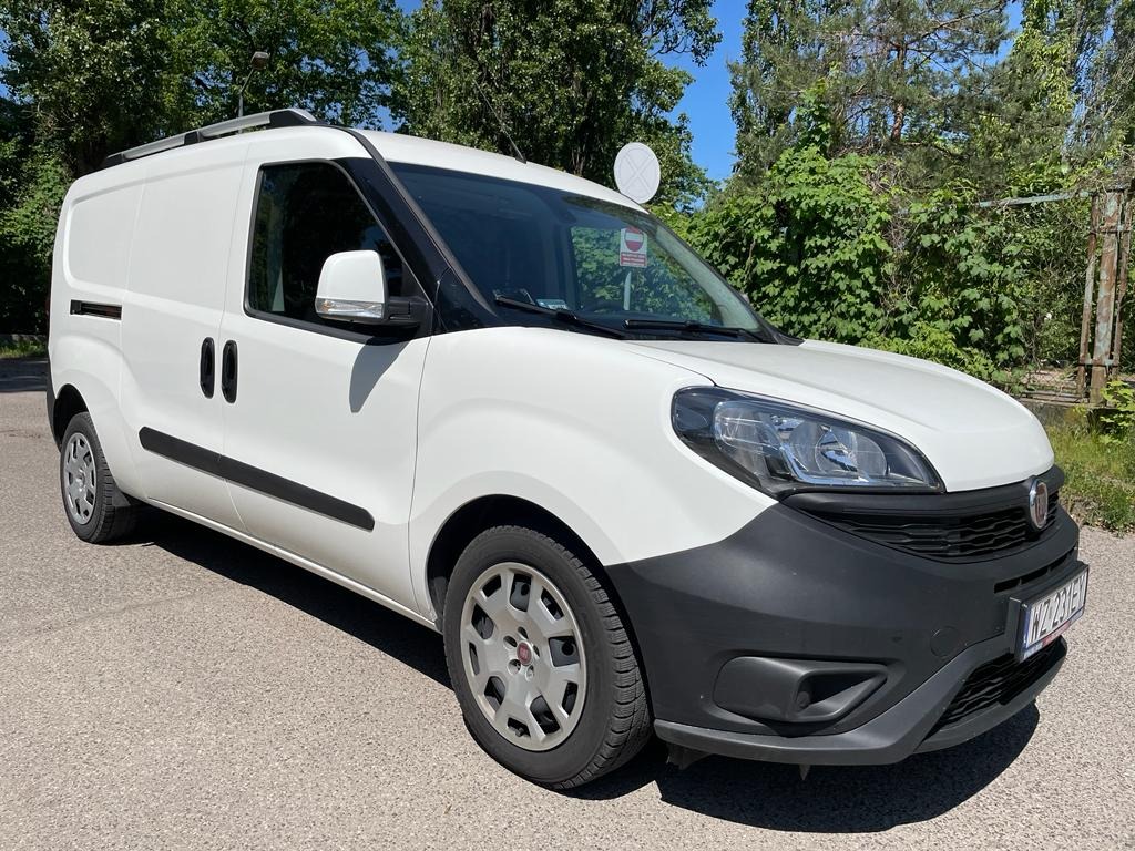 Biały samochód dostawczy Fiat Doblo z relingami dachowymi, zaparkowany na asfalcie, z widoczną tablicą rejestracyjną i drzewami w tle.