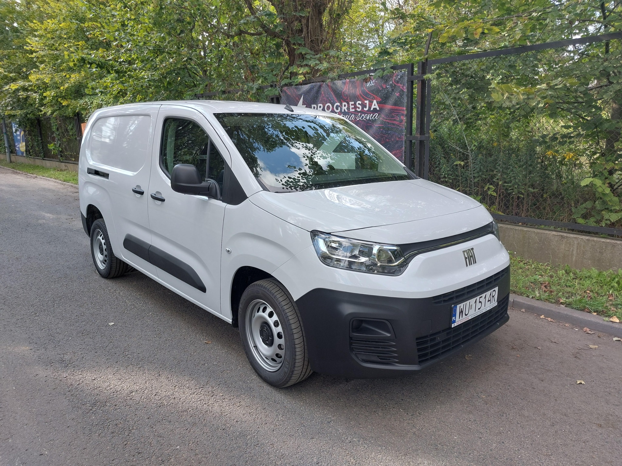 Biały samochód dostawczy Fiat na tle zieleni miejskiej i ogrodzenia z plakatem.