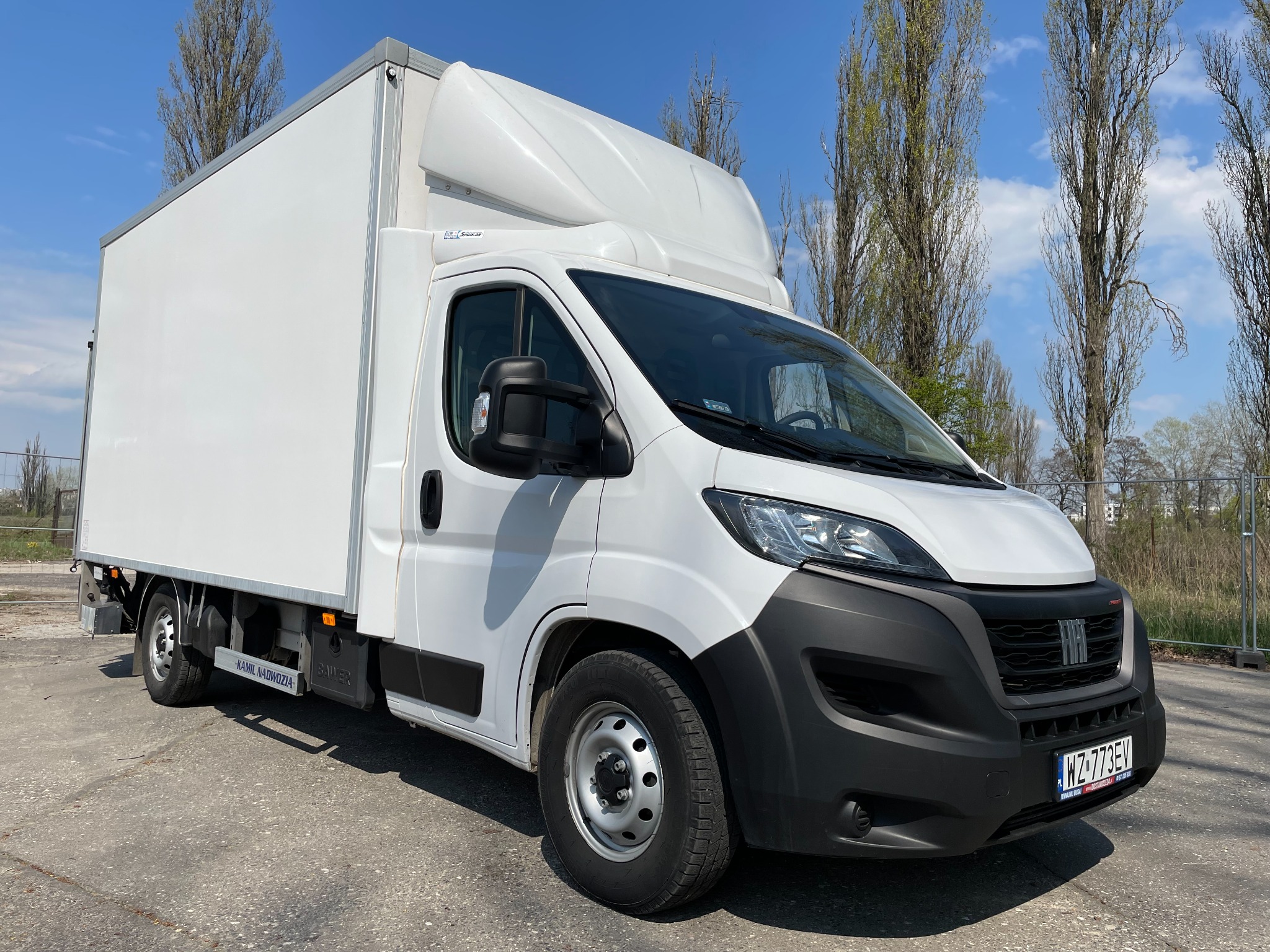 Biały samochód dostawczy Fiat Ducato z zabudową kontenerową, zaparkowany na betonowym placu w słoneczny dzień.