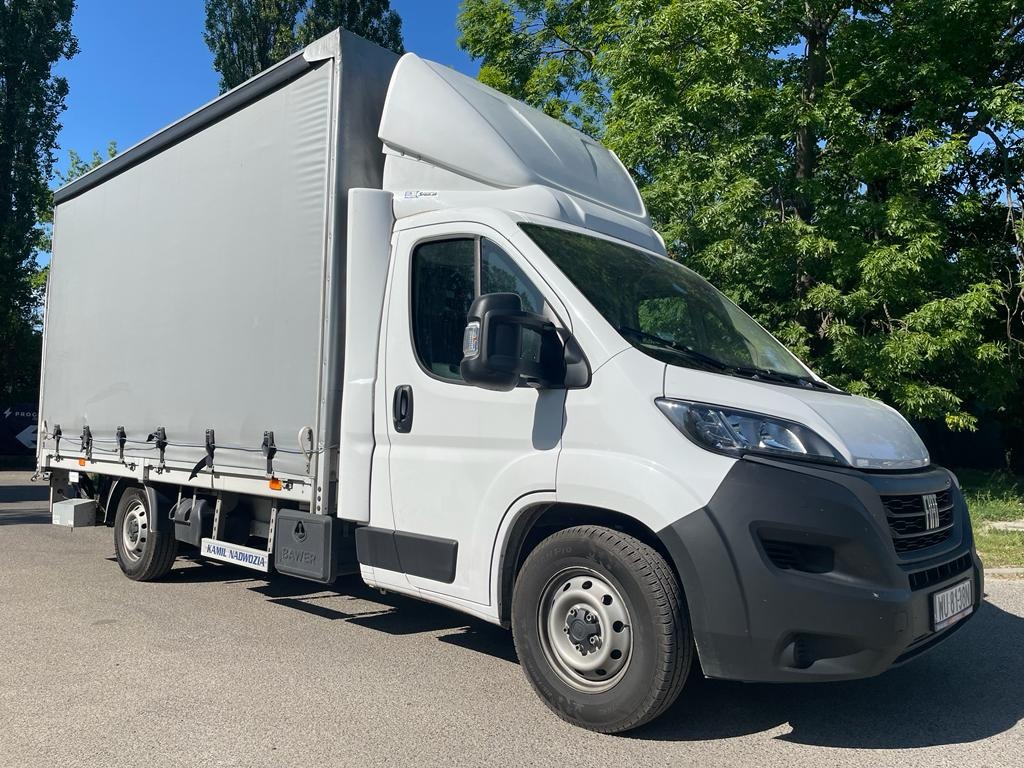 Biały samochód dostawczy z plandeką na parkingu w słoneczny dzień, widoczne logo firmy na boku.