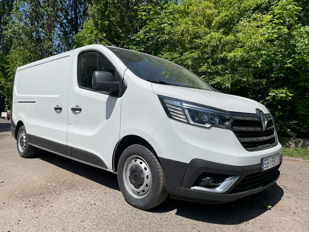 Biały samochód dostawczy Renault Trafic zaparkowany na szutrowej drodze, z widoczną tablicą rejestracyjną i zielonym tłem drzew.