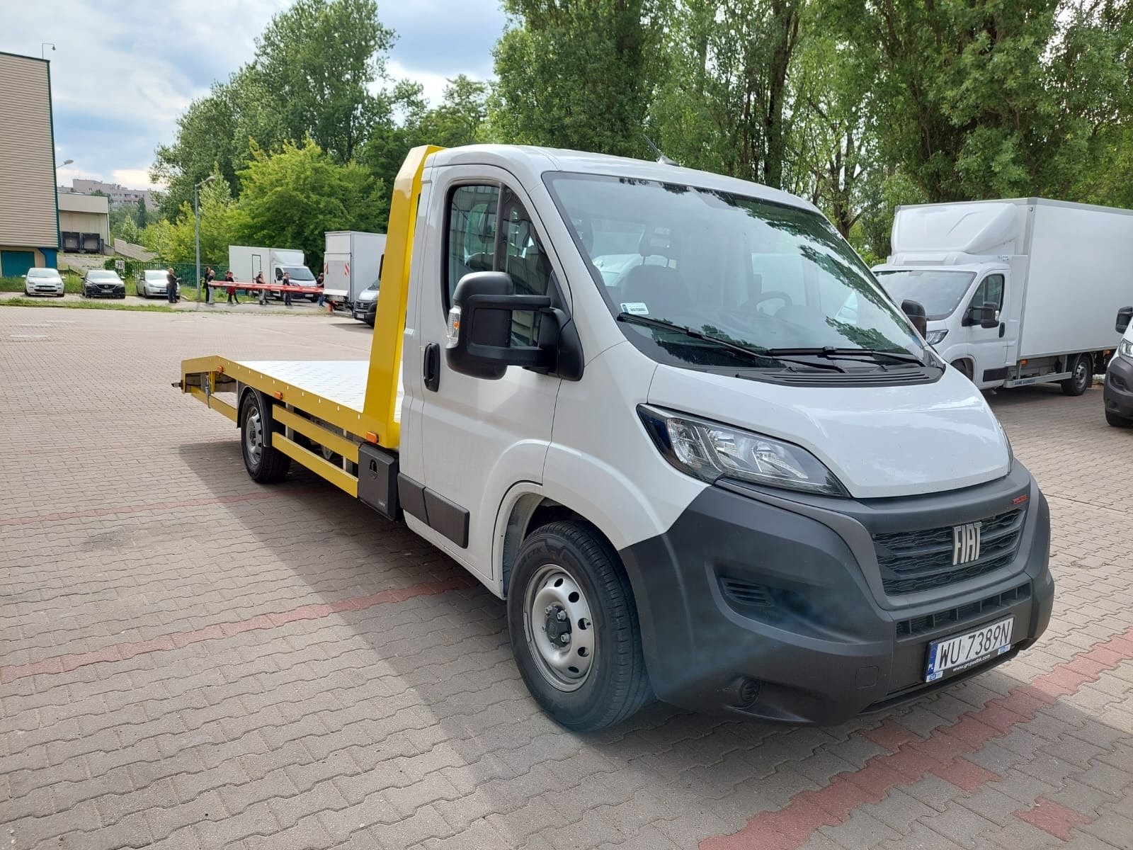 Biała laweta z żółtą platformą załadowczą na parkingu z kostki brukowej, widok z przodu i boku, kilka innych pojazdów w tle.
