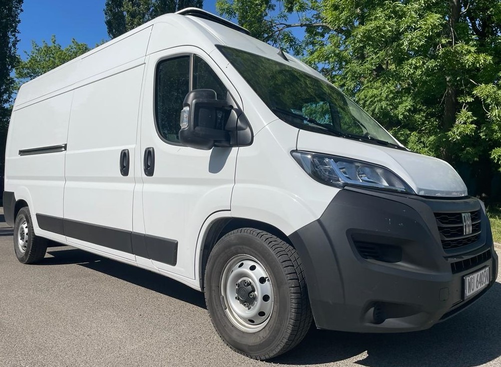 Biały samochód dostawczy Fiat Ducato zaparkowany na asfalcie w słoneczny dzień, widok z boku i lekko z dołu, z zielonymi drzewami w tle.