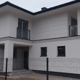 Nowoczesny, dwukondygnacyjny dom z białymi elewacjami i czarnym dachem, widoczny zza metalowego ogrodzenia, z elementami wykończeniowymi i pracownikami w tle.