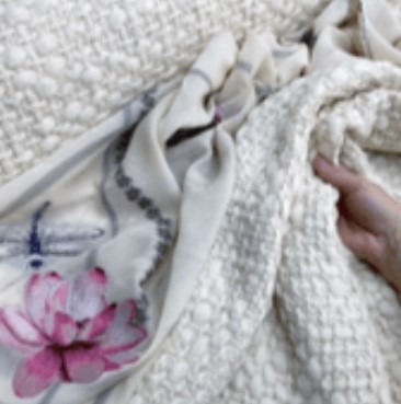 Dłoń układa warstwy tkanin: gruba, kremowa dzianina o wyraźnej fakturze, oraz delikatny materiał w odcieniach bieli z nadrukiem różowego kwiatu lotosu i szarych ważek.