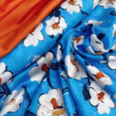 Błękitny materiał z nadrukiem białych kwiatów z pomarańczowymi środkami, częściowo zasłonięty pomarańczowym materiałem w lewym górnym rogu.