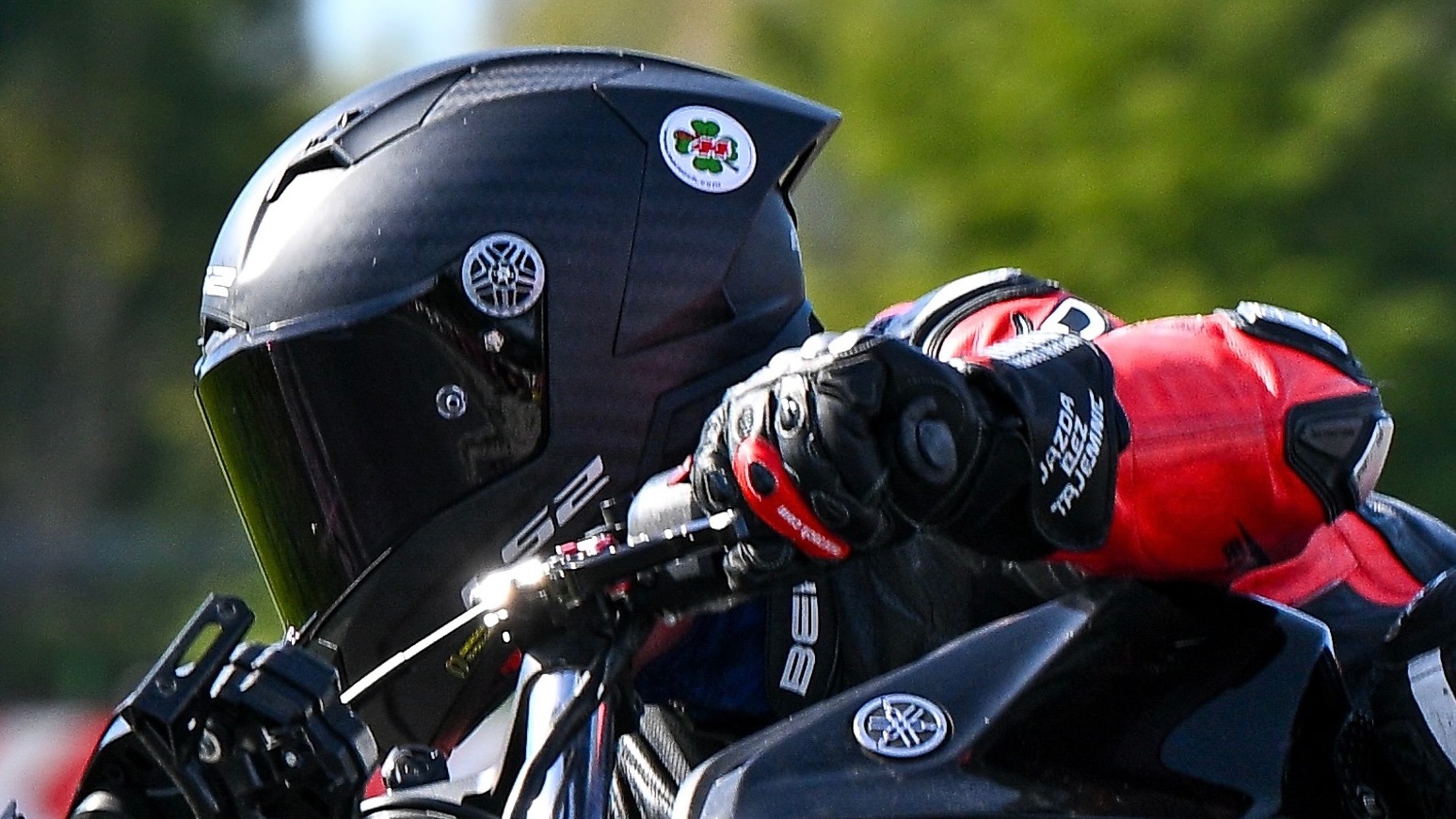 Motocyklista w kasku i rękawicach, trzymający kierownicę Yamahy. Na kasku naklejka z czterolistną koniczyną. Widoczne logo Yamaha na motocyklu.