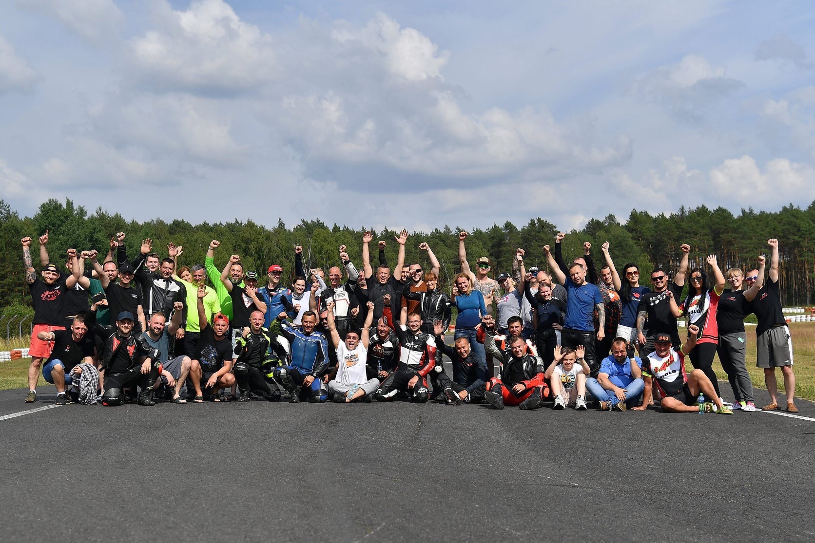 Grupa kursantów szkoły jazdy, w kombinezonach motocyklowych, radośnie pozuje na torze, z podniesionymi rękami, świętując ukończenie szkolenia.