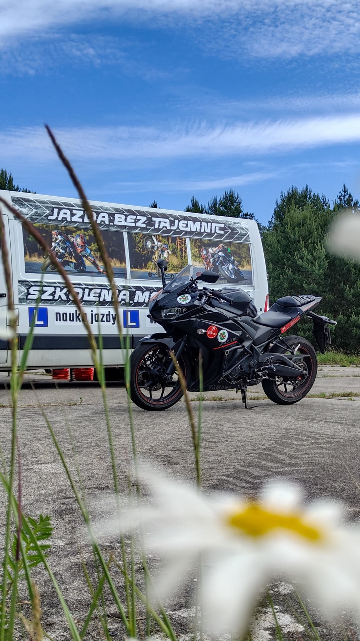 Czarny motocykl Yamaha na tle busa szkoły jazdy z napisem 'Jazda Bez Tajemnic', z kwiatami na pierwszym planie, w słoneczny dzień.