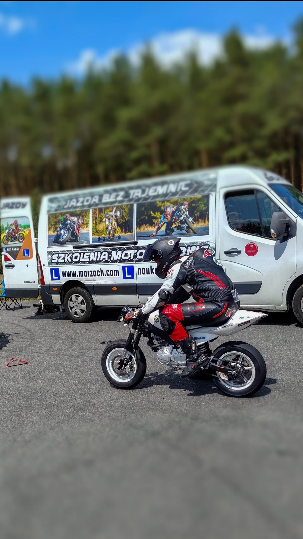 Kursant w pełnym stroju motocyklowym na mini motocyklu, w tle bus z reklamą szkoły jazdy 'Jazda bez tajemnic' i logo 'Szkolenia Moto'.