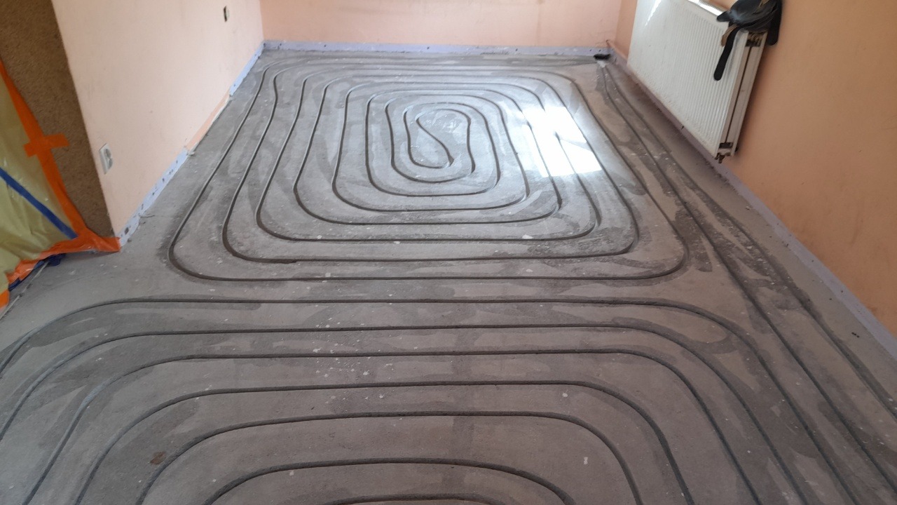 Węże ogrzewania podłogowego zatopione w szarej wylewce, układane spiralnie na podłodze w pomieszczeniu z pomalowanymi na brzoskwiniowo ścianami, widoczny fragment grzejnika.