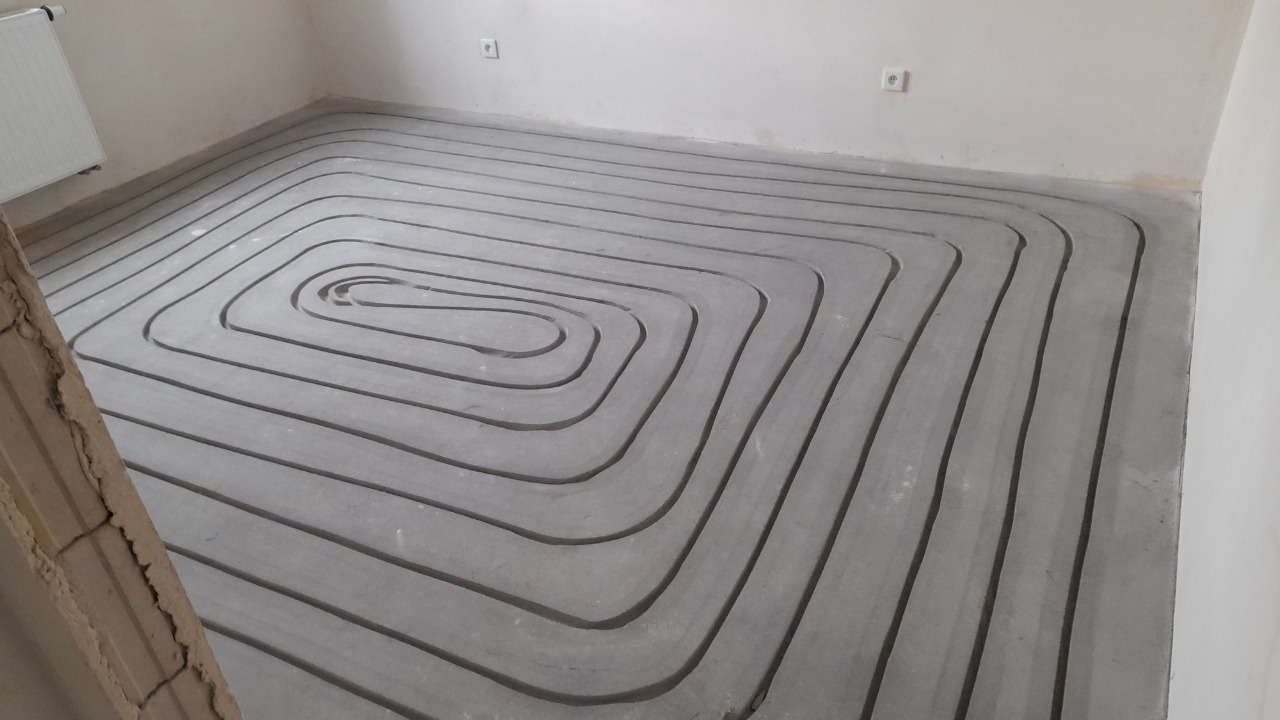 Szare rurki ułożone spiralnie w wylewce betonowej, przygotowane do systemu ogrzewania podłogowego, widoczne fragmenty ściany i grzejnika.