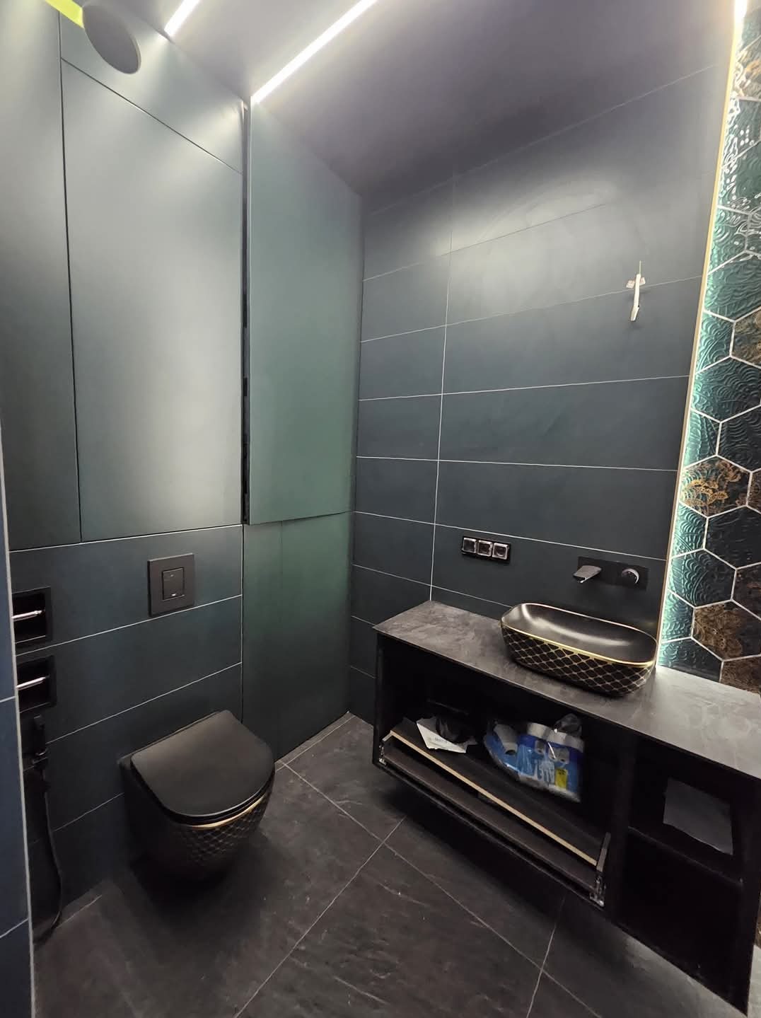 Elegancka łazienka w ciemnych tonacjach z czarną armaturą i dekoracyjną mozaiką. Nowoczesny design z podświetleniem LED i geometrycznymi wzorami na płytkach.