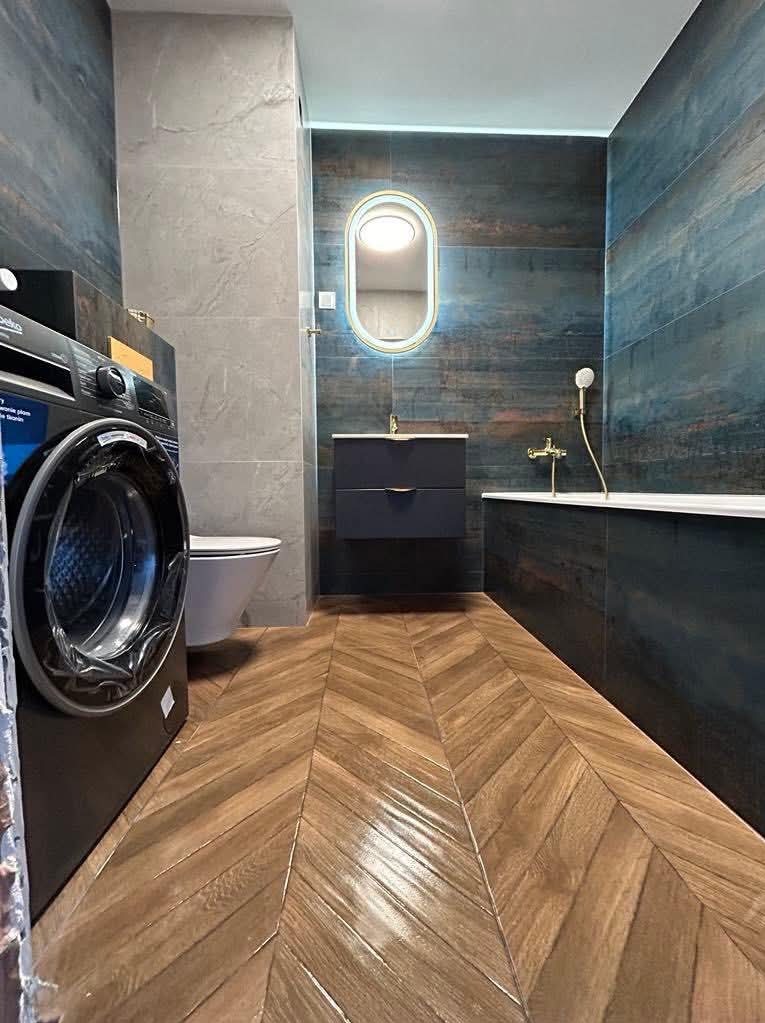 Nowoczesna łazienka z drewnopodobnymi płytkami ułożonymi w jodełkę, ciemnymi płytkami na ścianach i podświetlanym lustrem. Widoczna pralka i toaleta.