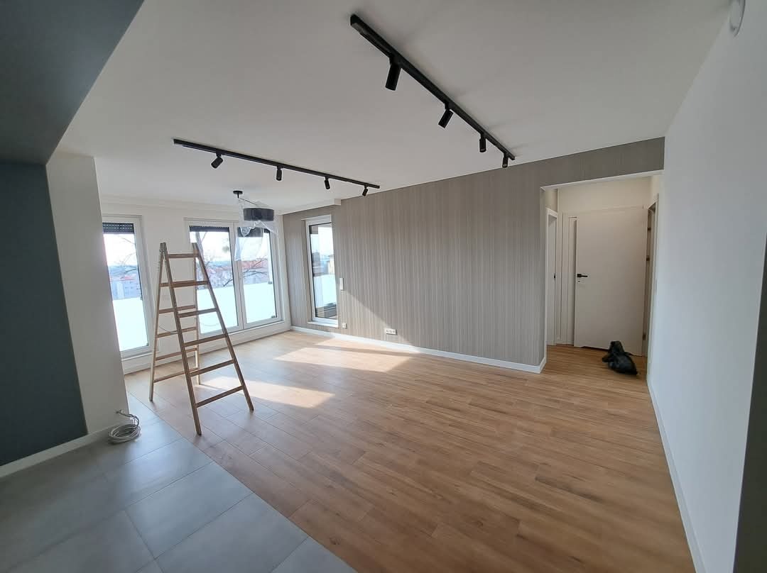 Jasne, przestronne wnętrze po remoncie: drewniana podłoga, szara tapeta, czarne lampy sufitowe. Aluminiowa drabina stoi przy oknie. Częściowo widoczne płytki podłogowe.