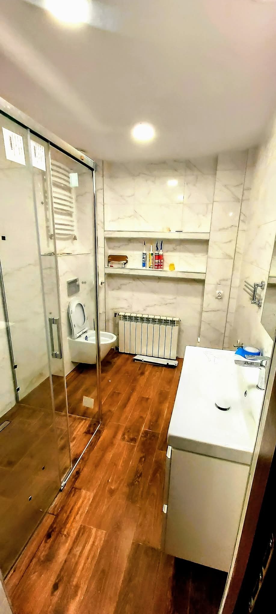 Jasna łazienka z kabiną prysznicową, drewnianą podłogą i marmurowymi płytkami. Widoczne: toaleta, umywalka z szafką, grzejnik i półki z akcesoriami.