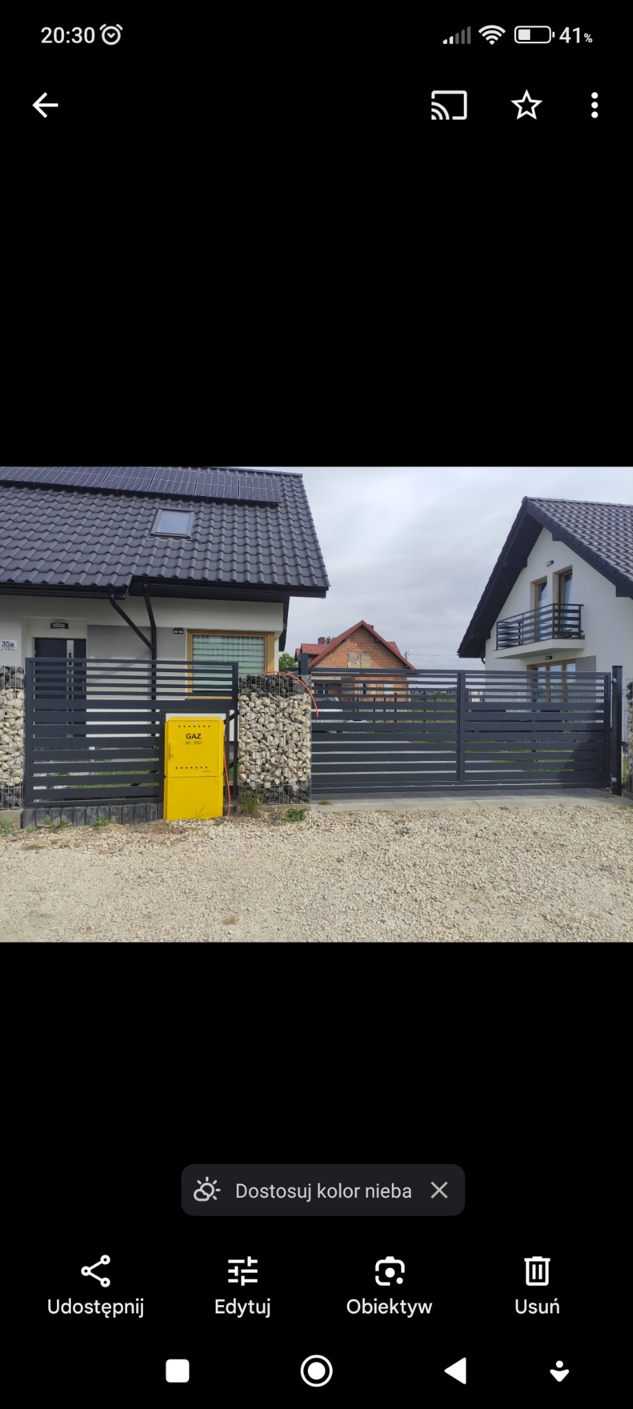 Nowoczesna, przesuwna brama wjazdowa w kolorze antracytowym, z poziomymi panelami, zamontowana przed domem z elewacją wykończoną kamieniem w koszach gabionowych, skrzynka gazowa w jaskrawym żółtym...