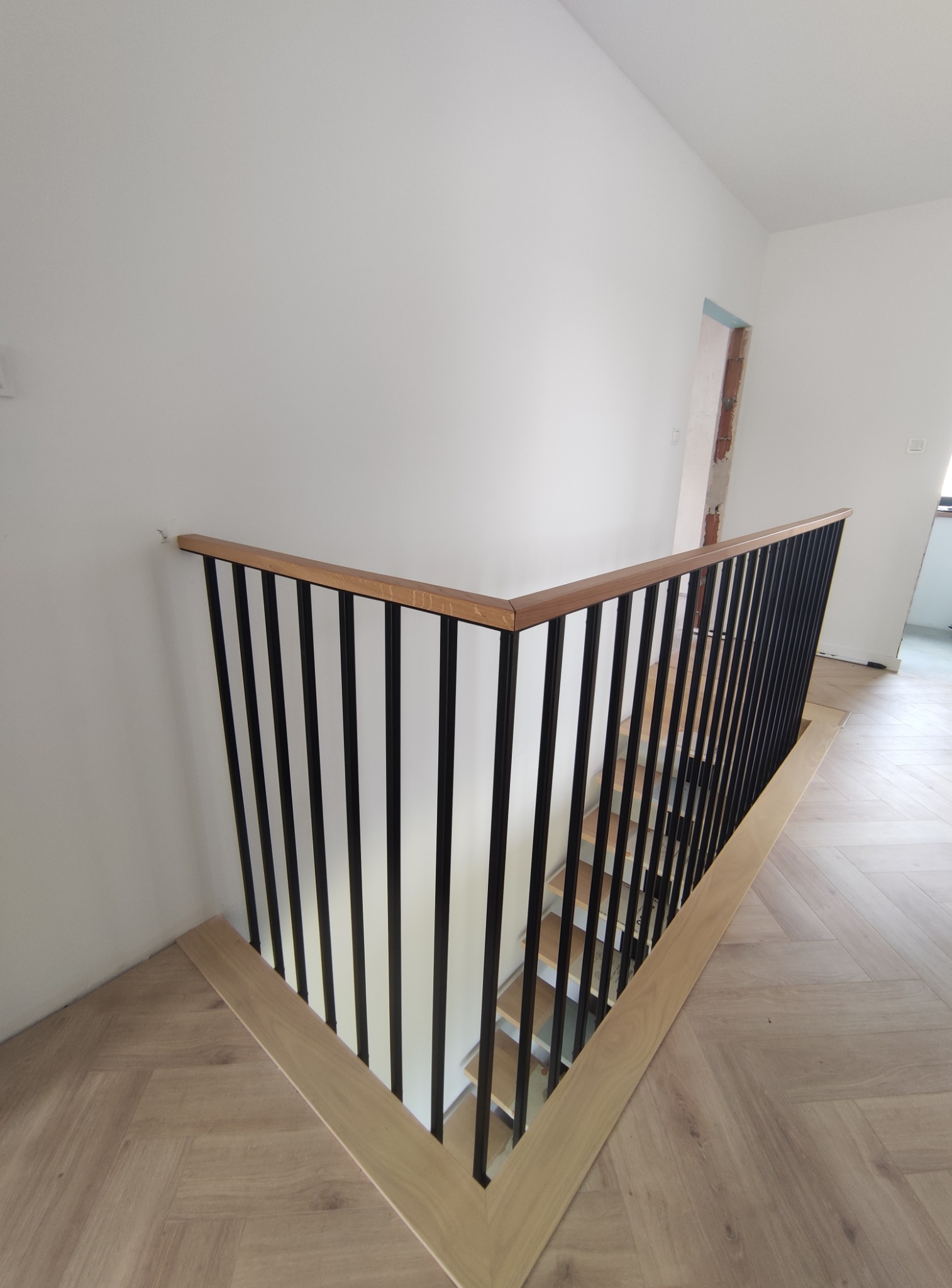 Nowoczesna balustrada schodowa z czarnymi, pionowymi prętami i drewnianym pochwytem, zamontowana na jasnych schodach wykończonych parkietem w jodełkę.