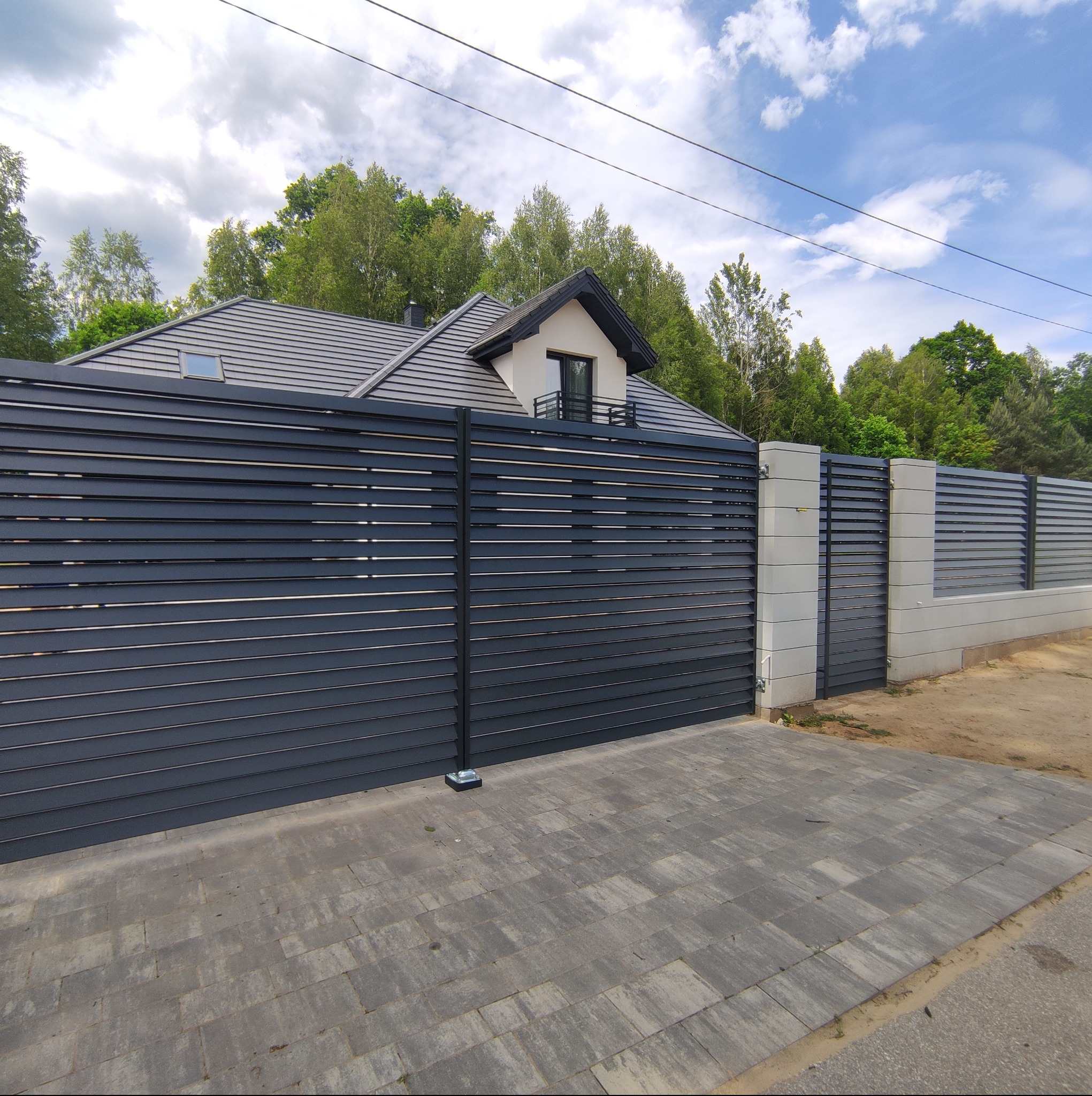 Nowoczesna, przesuwna brama wjazdowa z poziomych, grafitowych paneli aluminiowych, z widocznym fragmentem ogrodzenia o tej samej stylistyce i kostką brukową przed posesją, na tle domu i drzew.