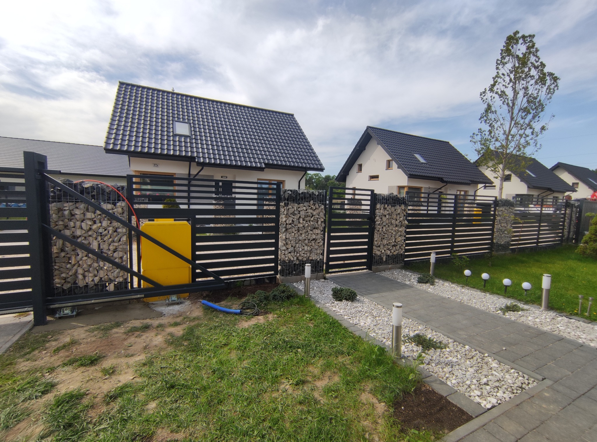 Nowoczesna posesja z przesuwną bramą wjazdową i ogrodzeniem z elementami gabionowymi, widoczne domy w tle.