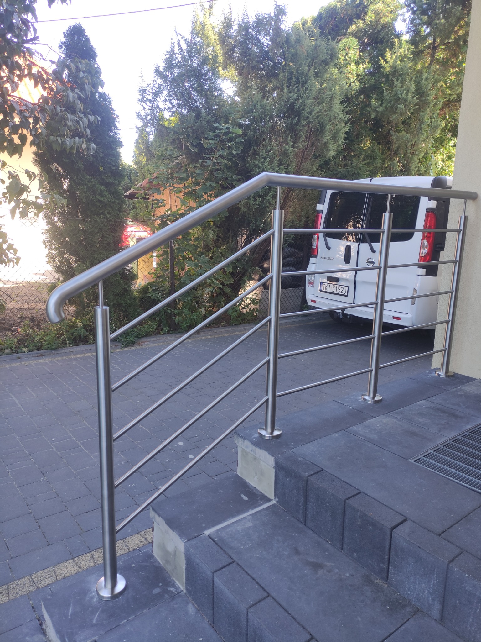 Nowoczesna balustrada ze stali nierdzewnej z poziomymi prętami, zamontowana przy schodach wejściowych do domu, z białym samochodem dostawczym w tle.