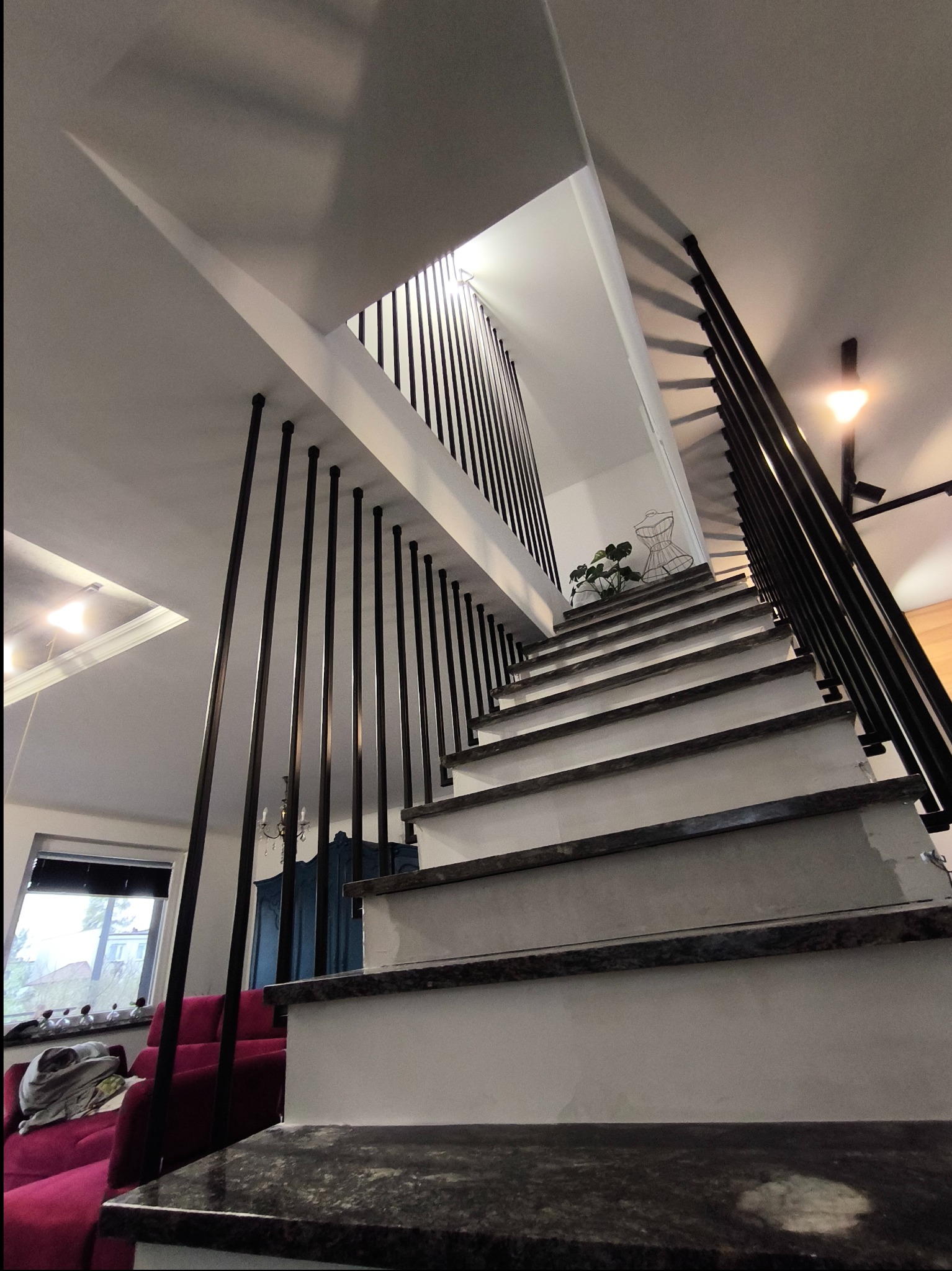 Nowoczesne schody wewnętrzne z czarną balustradą z prętów i stopniami z ciemnego kamienia, widok od dołu ku górze, w tle salon z czerwoną sofą i oknem.
