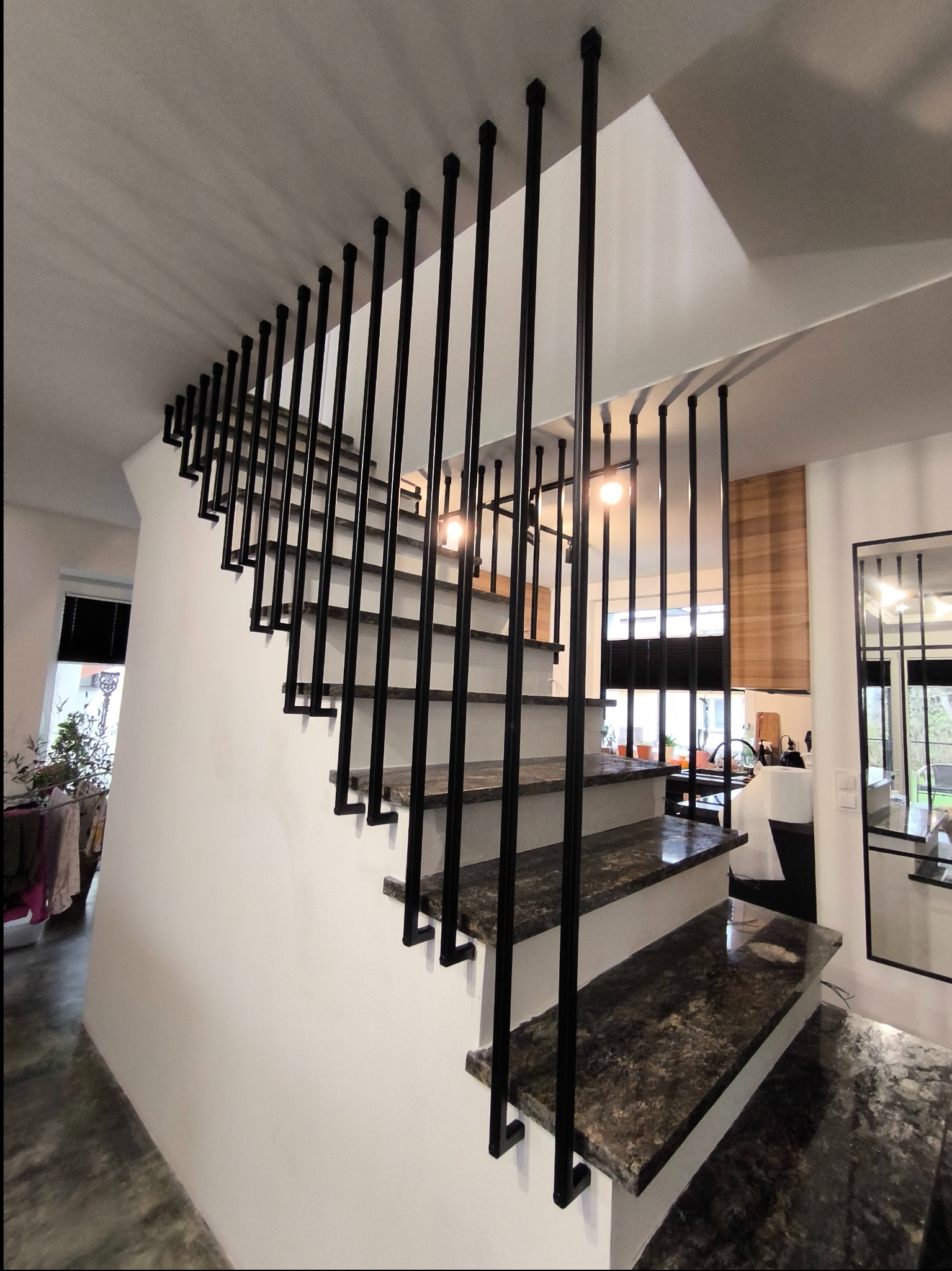 Nowoczesne schody wewnętrzne z ciemnymi, kamiennymi stopniami i minimalistyczną, czarną balustradą z pionowych prętów.