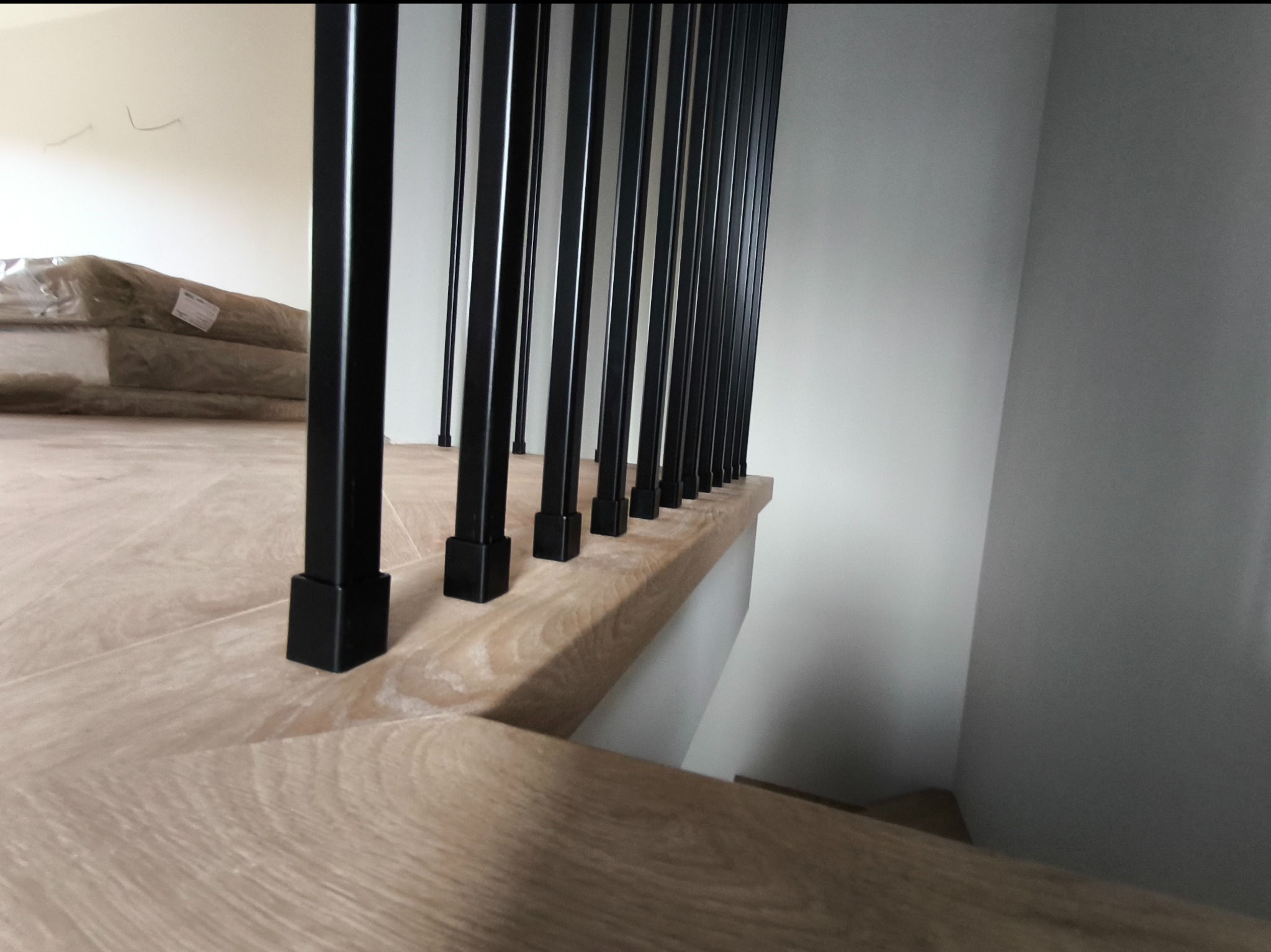 Fragment schodów z drewnianymi stopniami i czarnymi, pionowymi elementami balustrady, widok z góry na dół, w tle częściowo widoczne materiały budowlane zabezpieczone folią.