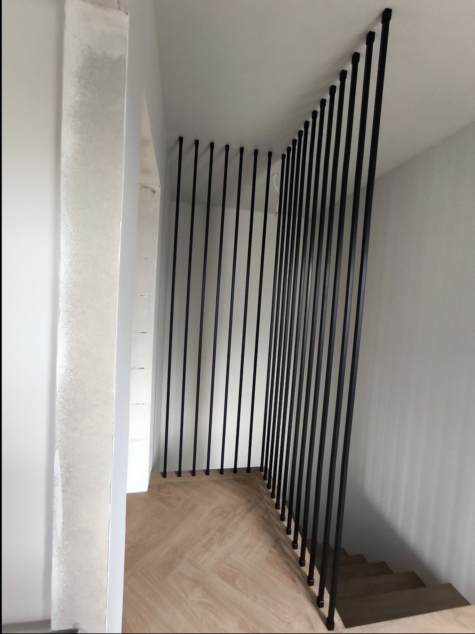 Nowoczesna barierka schodowa wykonana z pionowych, czarnych, metalowych prętów zamocowanych do sufitu i podłogi, z drewnianą podłogą w jodełkę i surowymi ścianami w tle.