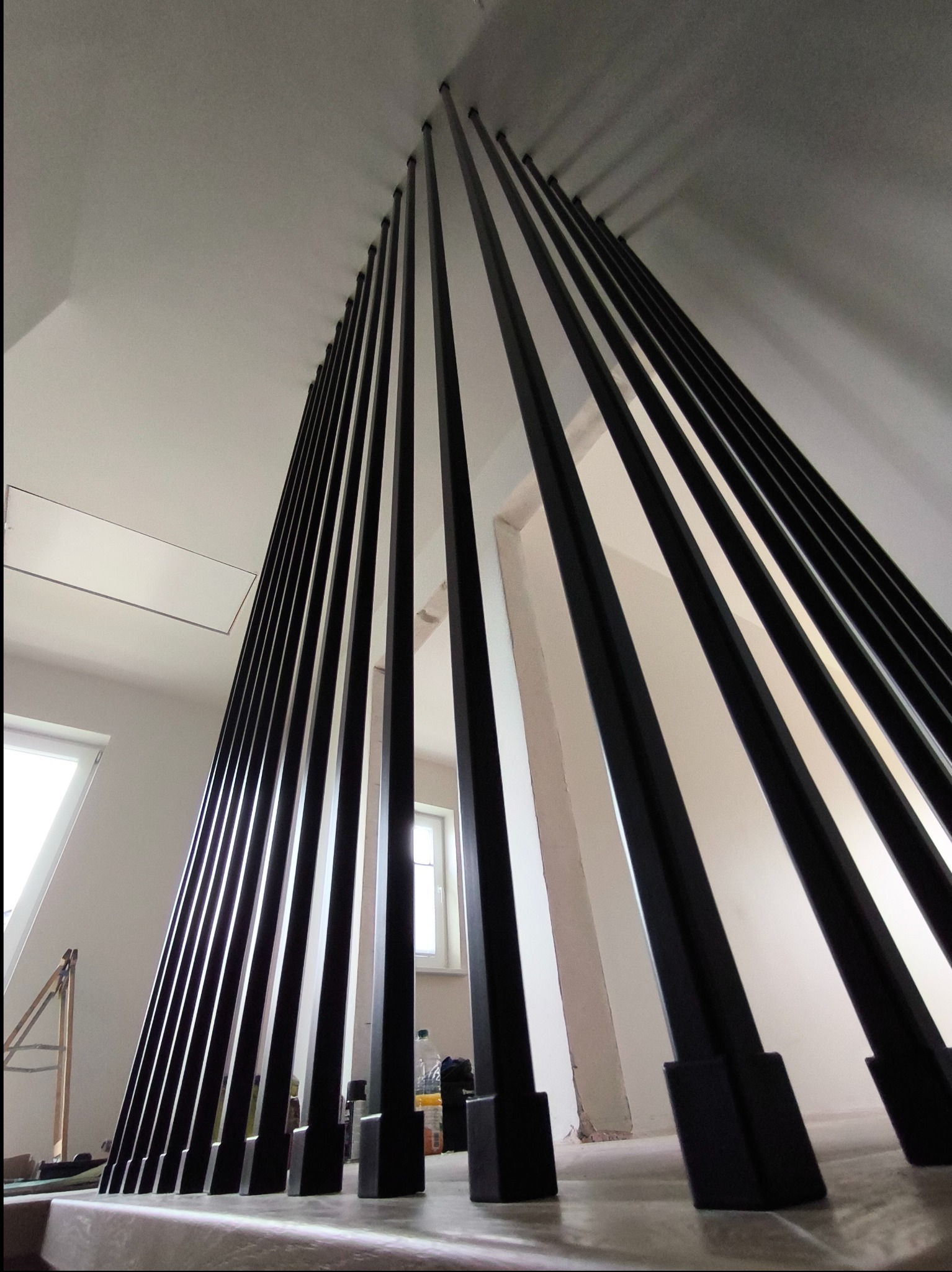 Nowoczesna, czarna barierka schodowa z pionowych profili, widok od dołu, zarys okna w tle.