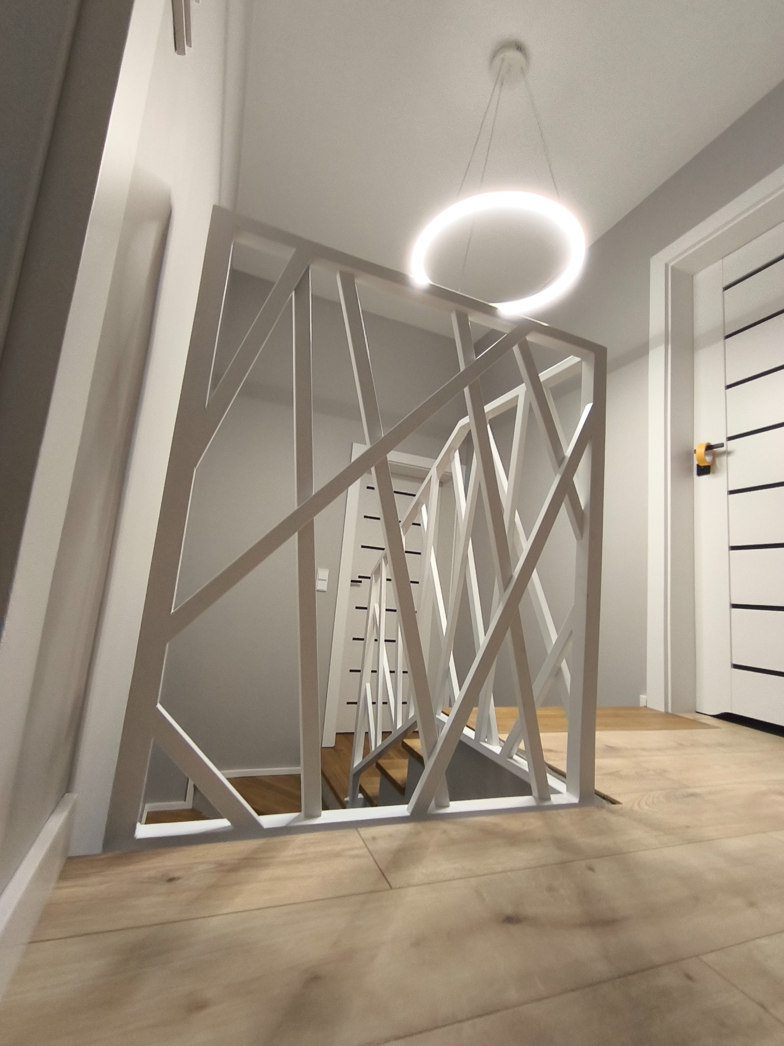 Nowoczesna, biała balustrada schodowa z geometrycznym wzorem, oświetlona okrągłą lampą wiszącą, widok z dołu na drewnianą podłogę.